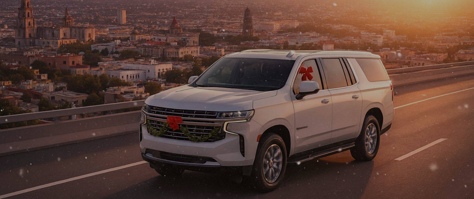 Camioneta Chevrolet Suburban 2025 blanca decorada con motivos navideños, circulando por un puente elevado con vista panorámica de Guadalajara al atardecer, con nieve ligera y la Catedral de Guadalajara al fondo.